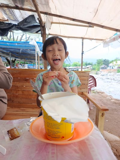 The image depicts a young girl, likely of Southeast Asian descent, in an outdoor, semi-covered setting in Meurah, Indonesia. She is positioned centrally, sticking out her tongue with her eyes closed, displaying a playful expression. Her short, dark hair has bangs, and she wears a light blue-green, short-sleeved top adorned with small patterns including rainbows, hearts, and butterflies. A gold-colored ring is on her right ring finger, and red marks are visible on her left forearm. In both hands, she holds a small, light-colored, spherical food item, possibly a dumpling or sweet.
To her immediate left, the shoulder and arm of an adult in a beige garment are partially visible. In the foreground, on a plastic-covered table, a large yellow cup (likely containing instant noodles or a drink) rests on an orange plastic saucer. Its white paper lid is partially peeled back, and a silver spoon is inserted. A small, dark-brown rectangular packet in clear plastic lies on the table to the left of the cup.
The girl is seated on a slatted wooden bench. Above, a makeshift canopy, constructed from light-colored tarpaulins and some black sheets, is supported by wooden beams. A black and white banner, partially obscured, hangs from the structure, with "BIG COFFEE" discernible.
The background reveals a wide, shallow, rocky riverbed with flowing water. On the opposite bank, several simple houses are nestled among trees, suggesting a village or rural settlement. Distant figures are visible near blue tarp structures further back on the left, and a solitary figure stands in the river. The overall environment is an informal riverside eating or resting area, under bright daylight.