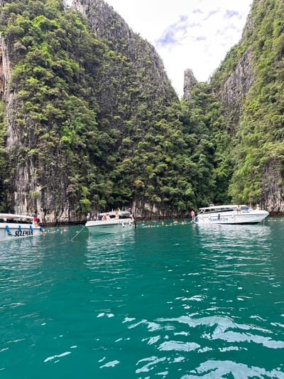 The image depicts a serene turquoise bay in Haad Yao, Thailand, flanked by towering limestone cliffs densely covered in lush green vegetation. Several speedboats are anchored in the calm water. On the left, a white speedboat with "SULEMAN" emblazoned on its side is docked. To its right, another white speedboat, identified as "Diamond Sea," is also moored. A group of people are visible on the deck of "Diamond Sea," and some are in the water, swimming or floating near orange buoys that mark a designated area. The water's surface is gently rippled, reflecting the overcast sky above.