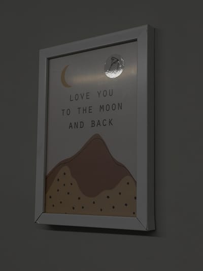 The image shows a framed print hanging on a wall. The print features a minimalist design with the words "Love you to the moon and back" written vertically, accompanied by a crescent moon and a stylized, abstract mountain shape with dotted details. A reflective sticker is affixed to the print. The frame is white. The wall has a neutral color. The location in the city of Zagarancea, Moldova, is not discernible.