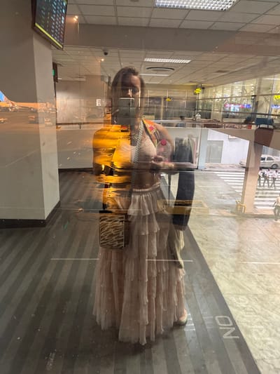 This media file shows a reflection of a woman standing in what appears to be an indoor, modern setting with large glass windows. The woman is wearing a light-colored, ruffled dress and a mask, and is holding a pink object and a bottle. She is looking towards the camera, and her reflection is visible in the glass.
The scene outside the glass appears to be a parking area with striped flooring and parked cars, suggesting a public space such as an airport or a shopping mall. There is also a sign visible in the reflection that reads "PARKING ON". The lighting suggests it is either dusk or nighttime.
The image's orientation is rotated, with the subjects appearing vertically. The overall atmosphere is a bit distorted due to the reflection, creating a sense of depth and layering. The environment seems clean and well-lit, with a mix of natural and artificial light sources.
The provided location context is Katunayake, Sri Lanka.