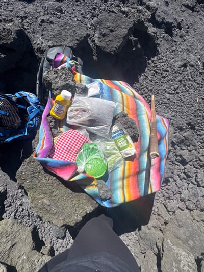 A colorful striped blanket sits on a rocky surface. It holds various items: a wooden stick, black rocks, a bottle of Hellman's mayonnaise, plastic bags (one with tortillas), wrapped limes, and a red and white checkered cloth. A blue backpack rests below the blanket. A leg wearing black pants is visible on the right, suggesting someone is nearby.
