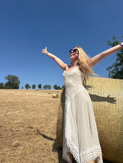A woman with blonde hair is standing in a field of hay bales under a clear blue sky. She is wearing a white lace dress and sunglasses, with her arms outstretched as if embracing the moment. The field appears to be in a rural setting, possibly in or around Tandel, Luxembourg, given the presence of hay bales and a sparse treeline in the background. The sun is shining brightly, casting a shadow of the woman and her outstretched arms on the hay bale beside her. The overall scene suggests a warm, sunny day in the countryside.