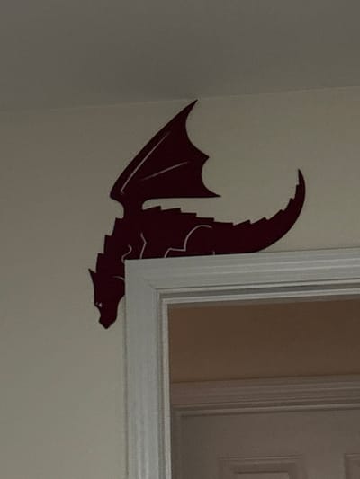 A maroon dragon wall decal is affixed to a light-colored wall adjacent to a white-framed window or door. The dragon, depicted in profile, exhibits textured scales along its curved neck and head. Its wing is partially obscured, but the distinct silhouette and serrated edges are visible. The dragon's head is close to the window frame, suggesting it is flying toward or beside it. The background behind the frame appears to be a light tan color.