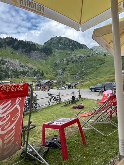 The image captures a detailed outdoor scene in Châtel, France, set at the base of a mountainous landscape under a partially cloudy sky. In the immediate foreground, two large yellow outdoor umbrellas provide shade, with the word "PELFORTH" visible in reverse on one. Directly below, a small red rectangular table rests on the grass, holding a black smartphone and black earbuds. Three red fabric deck chairs are positioned on the grass; the closest chair, partially obscuring the view, prominently displays the "Coca-Cola" logo. A green backpack with a "POC" logo is on the ground near this chair, and another dark backpack is situated under the red table.

Beyond this outdoor seating area, a wooden post-and-rail fence separates a gravel expanse. Approximately 10 to 15 individuals, many equipped with mountain bikes, are gathered on this gravel area. Several mountain bikes are propped against the fence, while one bike lies on its side on the grass next to the fence, accompanied by a red and black full-face helmet. A dark-colored pickup truck is parked on the gravel, near a wooden A-frame sign.

The background is dominated by steep, verdant mountain slopes covered in grass and scattered coniferous trees. Rocky outcrops and scree are visible on the mountainsides. A ski lift system, identified by its poles and cables, ascends one of the mountains towards a visible structure on a peak. Several traditional alpine chalet-style buildings are visible nestled in the mid-ground, at the foot of the mountains.