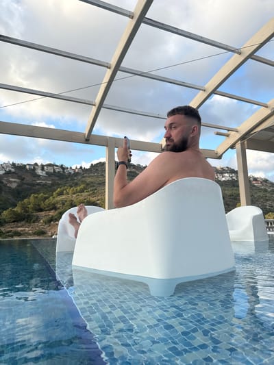 In Tirat Karmel, Israel, a shirtless man with short, dark hair and a beard sits in a white chair partially submerged in a swimming pool. He is holding a dark-colored smartphone in his right hand and looking away from the camera towards the right. His bare feet are visible resting on the bottom of the pool. The pool has a tiled bottom and the water reflects the cloudy sky and a wooden and metal structure overhead. In the background, a hillside is covered with green vegetation and dotted with white houses. Another white chair is visible to the right of the man.