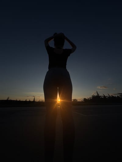 A photograph taken in Kipra, Bulgaria shows a silhouetted woman standing on a road as the sun sets. She has her hands behind her head. The sky is darker blue above her and closer to the horizon, the sky is lighter with some scattered clouds. Silhouetted trees are visible on the horizon.