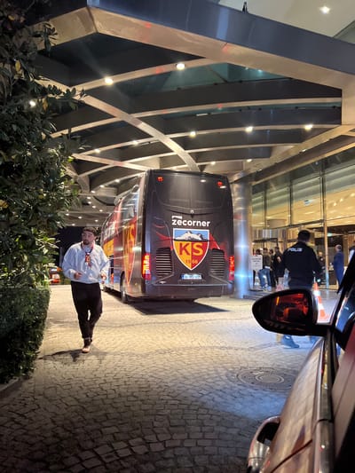 This image depicts an outdoor scene at the illuminated entrance of a modern building, likely a hotel, in Bağcılar, Turkey, observed during nighttime.
In the midground, a dark-colored coach bus, bearing "Zecorner" branding and a "KS 1966" logo with a red and yellow crest on its rear, is positioned facing the building entrance. Its license plate, "38 KS 835," is clearly visible. The bus features red and yellow graphic accents along its side.
To the left of the bus, a male individual with a beard, wearing a light-colored long-sleeved top (possibly a white sweatshirt), dark trousers, and light-colored sneakers, is captured in motion, moving from right to left across the cobblestone pavement. He appears to be jogging or walking briskly.
In the background, behind the bus, the brightly lit glass facade and entrance of the building are visible. A male individual in a dark uniform with "POLIS" emblazoned on his back stands facing the entrance, accompanied by several other indistinct figures congregated near the automatic glass doors. The entrance is covered by a geometrically structured canopy with numerous embedded downlights.
In the immediate foreground, to the right, the front-right section of a red vehicle is visible, including a portion of its fender, headlight, and side mirror, from which the photograph appears to have been taken. Two orange traffic cones are situated near this vehicle, one partially reflected in the side mirror. Green foliage is visible on the far left, bordering the cobblestone path.
