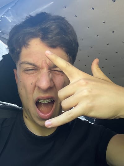 The image shows a close-up selfie of a young person with braces, who is winking and sticking out their tongue, making a rock-and-roll hand gesture. They are wearing a black t-shirt. The background appears to be the interior of a vehicle, possibly a car or van, with a grey headliner adorned with small, reflective dots. The lighting suggests an indoor environment, possibly during the day. There is no visible indication of the city of Taran, Bulgaria, in the image.