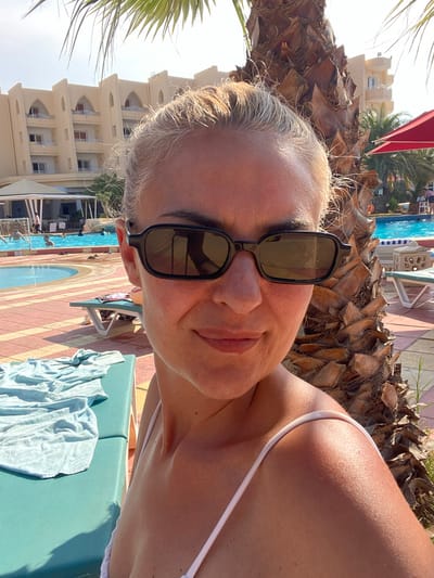 A woman wearing sunglasses and a white swimsuit is photographed on a lounger near a swimming pool at a resort in Monastir, Tunisia. The pool is surrounded by a red and yellow patterned tiled area, palm trees, and a hotel building in the background. Blue towels are on the lounger.