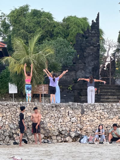 In Kuta Selatan, Indonesia, a group of people are gathered on a sandy beach in front of a stone wall and a weathered stone structure. Three women are engaged in what appears to be a yoga or meditation session on top of the wall, with their arms outstretched. Behind them, a dense canopy of trees and a palm frond are visible, framing the scene. To the right of the stone structure, there are steps leading up to an entrance. On the beach, two young men are conversing, one shirtless and the other wearing a black t-shirt and shorts. Further to the right, a seated woman and man are also present on the sand, with several pairs of footwear and bags nearby. The overall setting suggests a relaxed, outdoor activity taking place in a coastal environment.