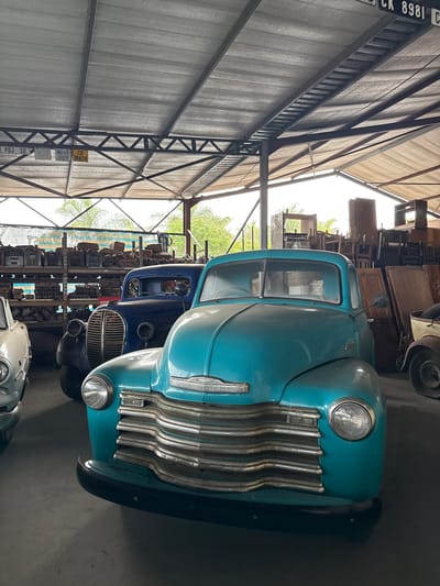 The image shows a warehouse or garage setting in Kraaifontein, South Africa, housing a collection of vintage vehicles. Prominently featured is a light blue Chevrolet pickup truck, with its front grill and headlights clearly visible. Behind the blue truck, a dark blue truck of similar vintage is partially visible. In the background, shelves are filled with various automotive parts and other items. The structure of the building includes metal support beams and a corrugated metal roof. Several vehicle registration plates are affixed to a support beam.