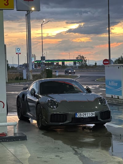 A dark grey Porsche 911 Turbo S is parked at a gas station in Drazhevo, Bulgaria. The car is positioned on a wet concrete surface, reflecting the surrounding lights and the vibrant sunset sky. The sky is filled with dramatic clouds illuminated by the setting sun, casting warm orange and yellow hues.

In the background, a highway overpass is visible with passing cars. A gas station sign with "OMV" branding is present, as well as an "AdBlue" dispensing unit. The overall environment suggests an evening scene at a roadside service area. The car's license plate reads "CB 0911 OH". There are no people visible in the image. The wet ground indicates recent rain or cleaning. The lighting suggests it is either dusk or dawn.