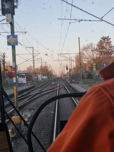 The image, taken from inside a train moving along a track in Rheinstetten, Germany, shows the railway lines stretching into the distance. The track is lined with overhead wires and support structures, signal lights, and signs. A railway platform is visible on the right side of the frame, with markers indicating platform number and information. An orange sleeve or garment is prominent in the foreground on the right, obstructing the view slightly. A smartphone device is on the lower side, partially in focus. The sky is overcast and light, with trees and buildings lining the horizon. The red signal light indicates possible restrictions ahead for the train movement. Several parked trucks can be seen in the distance.