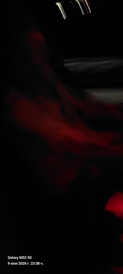 The image is a dimly lit, abstract photograph with a dark background and smudges of red light scattered across the frame. The lighting appears to be from an artificial source, possibly car headlights or streetlights, creating blurred streaks of light. In the bottom left corner of the image, there is text that reads "Galaxy M52 5G 9 юни 2026 г. 23:38 ч.", indicating the device used to capture the photo and the date and time it was taken. The location is identified as Amposta, Spain. There are no discernible people, subjects, or specific objects in the photograph due to the low light and abstract nature of the image. The overall scene is dark and indistinct, with the red light creating a somewhat dramatic or mysterious atmosphere.