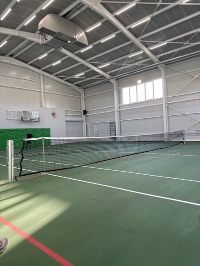 An interior view of a modern indoor sports facility in Gorna Mitropolia, Bulgaria, presents a tennis court with a green hard surface marked with white lines for tennis and additional red lines. A black tennis net is stretched across the court, secured by silver posts. One yellow tennis ball rests on the court beyond the net, while another is positioned mid-air near an adult male on the far side of the court. This individual, dressed in dark clothing, is partially obscured but appears to be holding a tennis racket, positioned as if to strike the ball.
The facility features high, gabled ceilings with exposed white metal trusses supporting numerous linear fluorescent lights and a large, curved silver duct system. The walls are constructed from white corrugated metal panels. On the left wall, a basketball hoop with a clear backboard and orange rim is mounted above a green padded wall section. A large grey roll-up door and a standard white door are visible on the far wall. A series of rectangular windows situated high on the right wall admit natural light. The floor shows subtle areas of discoloration and wear. The toe of a grey shoe is visible in the bottom left foreground, indicating the photographer's close proximity to the court.