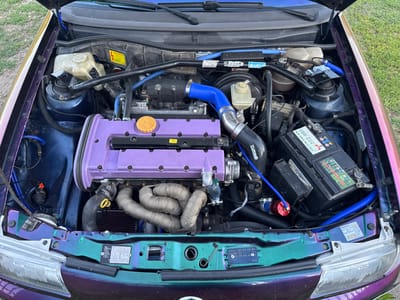 The image shows a modified car engine bay, likely taken outdoors in Lepitsa, Bulgaria. The engine has a prominent purple valve cover and braided exhaust manifold wraps. Various aftermarket performance parts are visible, including blue silicone hoses and what appears to be a turbocharger intake. The engine bay is clean and organized, with a visible battery and fluid reservoirs. The car's exterior, visible at the edges, has a multi-colored iridescent paint job. The surrounding environment is grassy, suggesting a residential or outdoor setting. There are no people present in the image.