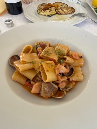 The image depicts a meal served on a white tablecloth within a restaurant environment in Nardò, Italy. The specific establishment or precise street location within Nardò is not ascertainable from the visual content.

In the foreground, a large white bowl contains a serving of paccheri pasta. The golden-yellow pasta pieces are coated in an orange-hued sauce. Visible ingredients include multiple clams, some in opened shells and some exposed, and lighter-colored protein pieces consistent with cooked squid or fish. The dish is garnished with finely chopped green herbs sprinkled across the surface.

In the mid-ground, a white plate holds remnants of grilled vegetables, specifically slices of zucchini and eggplant, exhibiting char marks from grilling. A small, irregular orange object rests on one eggplant slice. A silver dining fork is positioned on the table, partially resting on this plate. To the upper left, a dark-colored bottle, consistent with a wine bottle, is partially visible. Adjacent to it is a small, silver-colored, screw-top cap. A clear drinking glass is partially visible on the upper right, alongside a yellow object, consistent with a lemon or fruit wedge. No individuals are present in the scene.