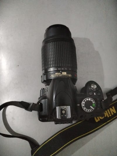 A black Nikon DSLR camera with a telephoto lens is placed on a light grey, textured surface, possibly a table or floor. The camera is angled diagonally, with the lens pointing upwards and to the left. The lens has zoom markings ranging from 55mm to 200mm. The camera body is black, and a black camera strap with yellow trim is visible, partially obscuring the right side of the camera. The camera strap has "Nikon" printed in yellow. A silver hot shoe mount is visible on top of the camera body. A mode dial is present on the top of the camera, showing various shooting modes including "AUTO" and "GUIDE". The background is a plain, light grey surface with some subtle texture and a shadow cast by the camera on the left side. There are no people or animals present in the image. The location is the city of Moara Domnească, Romania.