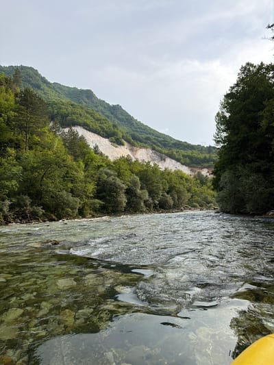 This is a landscape photograph taken from a body of water, likely a river or stream, with a bright yellow object visible in the upper right corner, possibly part of a kayak or raft. The camera is positioned low, close to the water's surface, looking towards a steep, tree-covered hillside. The water itself is clear, revealing a rocky bed beneath. The surface of the water is rippled, catching the light and creating a textured, dynamic appearance.
The hillside rises dramatically, densely populated with lush green trees and shrubs. A striking feature of the hillside is a large, pale, rocky scar running diagonally across its face, suggesting a landslide or quarry. Above the tree line, the sky is overcast and hazy, indicating a cloudy or misty day. The overall mood is serene and natural, capturing a moment of quiet observation in a wild, verdant environment. The location is identified as Polje Bijela, Bosnia and Herzegovina. There is no visible text in the image.