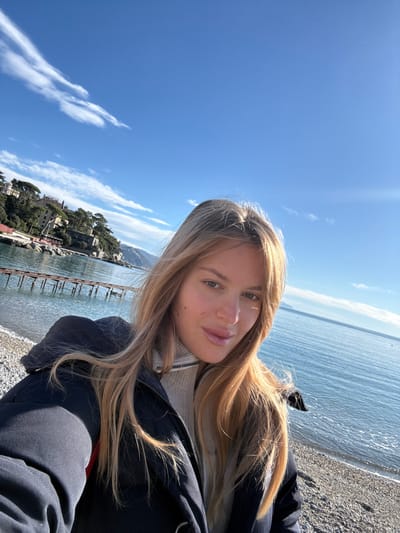 A young adult female with fair complexion and long, wavy blonde hair is captured in a selfie-style photograph. She is looking directly at the camera with a slight smile, wearing a dark-colored puffer jacket over a light-colored turtleneck sweater. The scene is set on a pebbled beach adjacent to calm, blue-green sea water. In the background to the left, a partially submerged wooden pier structure extends into the water. Behind the pier and beach, green, tree-covered hills rise, featuring several multi-story buildings with light facades and red-tiled roofs nestled amongst the vegetation. The right side of the frame shows the sea extending towards the horizon under a clear blue sky with sparse, wispy white clouds. Distant landmasses are faintly visible on the far right horizon. The scene is illuminated by bright, natural daylight.
This location is identified as the bay of San Michele di Pagana, a hamlet situated on the Ligurian coast of Italy, immediately adjacent to the city of Santa Margherita Ligure.