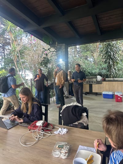 Under a wood-beamed structure, several people gather outside. A woman works on a laptop at a light-colored table cluttered with a purse, charger, and food containers. Other individuals stand in the background, some holding drinks, near a grill and coolers. Lush greenery is visible in the distance. Someone at the table eats plantain chips.