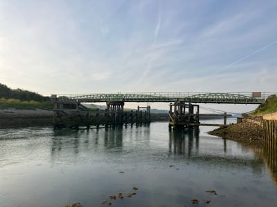 The image depicts a view of the old railway bridge over the River Ouse in Lewes, United Kingdom. The bridge is constructed with wooden pilings and a green steel superstructure. The river is calm with visible reflections of the bridge supports. The riverbanks are covered with vegetation and rocky embankments. In the background, there are power lines visible in the sky, and the sky itself is pale blue with scattered clouds.