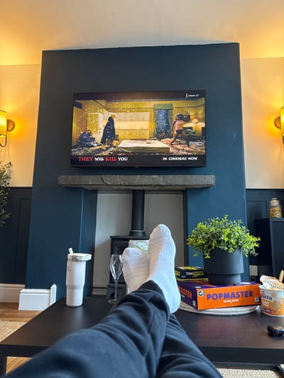 A person wearing white socks and a black sweater is watching the movie "They Will Kill You" on a television screen. The television is mounted on a wall above a decorative shelf holding a plant, glasses, and a white Stanley tumbler. On a nearby table are board games ("Pop Master", "Think Faster!"), a container of Golden Syrup, and a cup with a spoon. The wall is painted navy blue and has sconces above the television.
The location is not explicitly defined in the image.