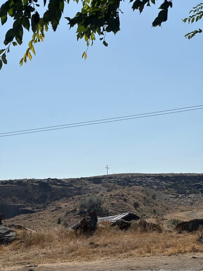 This image presents an arid, rocky landscape under a clear, light blue sky, within the city of Shirak, Armenia. Overhead, out-of-focus green leaves from a tree branch partially frame the upper portion of the scene. Two thin, dark horizontal utility lines traverse the sky in the upper mid-ground. The terrain consists of undulating, dry hills with scattered dark rock formations and sparse, golden-brown grasses. On the highest visible ridge, a solitary, dark-colored Christian cross stands upright. Below the ridge, in the mid-ground, a small structure with a dark, corrugated metal roof is visible amidst the dry vegetation and dark boulders. No individuals, animals, or active subjects are present.