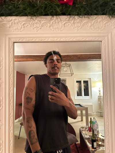 A person is taking a mirror selfie. They have tattoos on their arms and are exhaling smoke. The background shows a room with a table, wine bottles, and a window. The mirror is ornate with a white frame. A portion of a Christmas tree is visible in the lower left corner. The person is wearing a dark tank top and holding a phone in their hand.