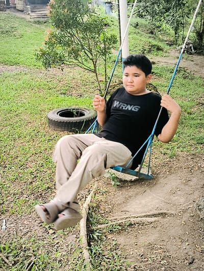 A young boy is captured in motion on a swing in an outdoor, green setting in Lambarotunong, Indonesia. The boy, who appears to be of elementary school age with dark hair, is positioned in the center-right of the frame, mid-swing. He wears a black short-sleeved t-shirt with the white text "WRNG" prominently displayed across the chest, and light tan or beige trousers. His feet, clad in sandals, are blurred due to the motion of the swing. His expression is neutral, looking slightly off-camera to the right.
The swing itself is simple, with blue-painted metal bars and white ropes or chains. The ground beneath the swing is a worn dirt path, surrounded by short, green grass interspersed with patches of dry leaves and exposed roots. To the boy's left, an old car tire lies on the grass, seemingly repurposed or discarded, with some small plant growth inside. Behind him and to his right, there are various trees and lush green foliage, indicating a natural, somewhat unkempt environment, possibly a park, backyard, or rural area. The lighting suggests it is daytime, with soft, diffused light, possibly overcast. In the background, hints of structures or buildings can be seen through the trees, further suggesting a semi-developed outdoor space.