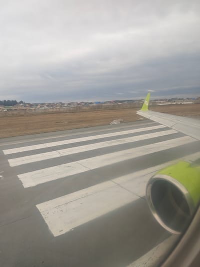 From the perspective of an airplane window, the scene depicts a landing strip in Vaniuki, Russia, marked with parallel white stripes. An S7 Airlines wing and engine are visible in the foreground. Beyond the runway is a flat, grassy field leading to a cluster of residential buildings. The sky is overcast with grey clouds.