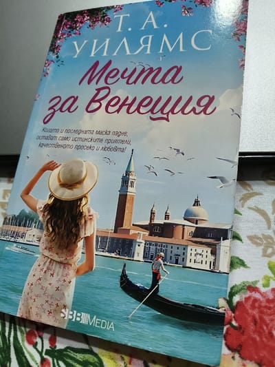 A photograph displays a paperback book cover, titled 'Т. А. УИЛЯМС Мечта за Венеция' (T.A. Williams, Dream for Venice) in Cyrillic, resting on a floral-patterned fabric surface. A plain, light-colored surface is visible in the upper right background. The book cover illustrates a vibrant scene of Venice, Italy. In the foreground of the cover, a woman with long wavy brown hair, wearing a straw hat and a light-colored, short-sleeved dress with a subtle red geometric pattern, is viewed from behind, looking out towards the water. Her right hand is raised, touching the brim of her hat. On the tranquil blue water, a black gondola is being propelled by a gondolier, dressed in a red-and-white striped shirt and a straw hat, using a long oar. The background of the cover image features iconic Venetian architecture, specifically the prominent Campanile di San Giorgio Maggiore and the Church of San Giorgio Maggiore, set against a clear blue sky dotted with numerous flying white seagulls. Pink floral motifs decorate the top border of the book cover, and the publisher's logo "BBBMEDIA" is present near the bottom. The depicted location on the book cover is Venice, Italy. The image itself provides no visual evidence to indicate that its physical capture location is in the city of Villa di Gemmano, Italy.