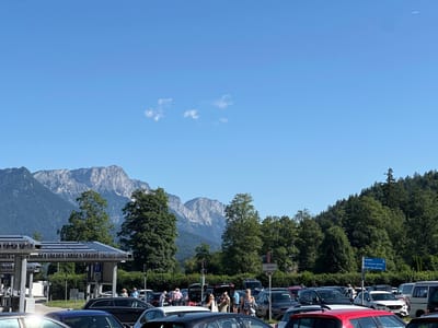 The image depicts a sunny outdoor scene at a parking lot in Schönau am Königssee, Germany. In the foreground, numerous cars are parked, with some people milling about. In the middle ground, there is a structure with solar panels on its roof, likely a covered parking area or charging station. Behind this, a line of lush green trees forms a backdrop, with towering, rugged mountains visible in the distance under a clear blue sky. The overall impression is that of a busy tourist area on a pleasant day.