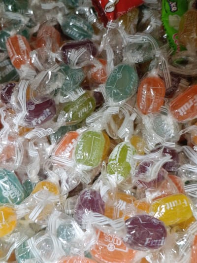 The image shows a close-up, overhead view of a pile of individually wrapped fruit candies. The candies are of various colors, including orange, green, yellow, and purple. Each candy is encased in clear plastic wrapping, tied at both ends with white twisted paper. Some of the wrappers have text and graphics visible, such as "Fruit" and "Bayan Sulu". The candies are jumbled together, filling the frame. There is no visible indication of a specific location in Martuni, Armenia, or any people, actions, or interactions. The environment is simply the collection of candies.