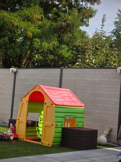 This image depicts a backyard scene in Grünstadt, Germany, on a sunny day. The focal point is a vibrant, multi-colored plastic playhouse with a red roof, yellow walls, and green accents. In front of the playhouse, a toy fire truck with a red cab and white body sits on a green lawn. To the right of the playhouse, a brown woven cube-shaped furniture piece and a decorative garden ornament of a rabbit leaning against a white volleyball are visible. A modern grey fence, possibly composite material, runs horizontally across the background, separating the yard from a dense green tree line. The ground in the foreground is paved with grey rectangular stones. The overall impression is of a well-maintained residential outdoor space, likely a backyard, designed for children's recreation.