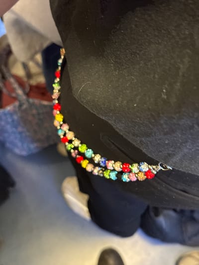 The image is a close-up, high-angle shot showcasing a vibrant, multi-colored beaded chain against a dark, possibly black, fabric surface. The chain is composed of numerous small, round beads, each appearing to be encrusted with rhinestones or crystals. These beads display a variety of bright colors, including red, yellow, green, blue, pink, and white/clear, arranged in an alternating pattern. A small silver-colored metal clasp is visible at one end of the chain, suggesting its use as an accessory like a bag charm, keychain, or decorative embellishment.
The dark fabric, which takes up a significant portion of the foreground and midground, shows some texture and minor lint or dust particles. The background elements are heavily blurred due to a shallow depth of field, rendering them indistinct. These blurry shapes and colors suggest an indoor setting, possibly showing other pieces of clothing, furniture, or floor, but no specific details can be identified.
No particular activity is taking place; the image is a static capture focusing on the accessory. The time of day is not discernible, but the lighting appears to be ambient. There is no visible text in the image. The scene is located in Bourg-la-Reine, France, and depicts a simple, detail-oriented view of a colorful item.