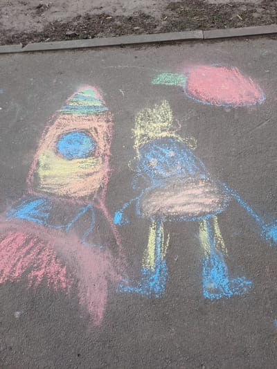 The image shows chalk drawings on asphalt pavement in a city location that may be in Меткомелино, Russia. The drawings include a rocket with a circular window, possibly flames emitting from the base, an object resembling a person or figure in space gear, and a shape that may be a flower or other abstract design. The drawings are executed in various colors of chalk including pink, blue, yellow, and green. The pavement is gray and shows typical wear and tear with an adjacent curb visible on the left side of the frame. The area near the curb has some loose dirt and debris. There are no people directly visible within the frame, but it may be assumed they are nearby if they drew on the ground.