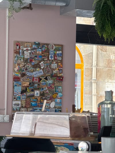 The image depicts an interior scene, likely a cafe or shop, with a view towards a wall and an exterior window.
On the left, a pale pink wall features a large, rectangular wooden board densely covered with approximately 100-150 diverse travel souvenir magnets. Notable magnet designs include place names such as "TORINO," "LOS ANGELES," "NEW YORK," "ISTANBUL," "JAMAICA," "CANADA," "MALTA," "ARGENTINA," and "STRASBOURG," alongside depictions of landmarks, flags, and cultural motifs. Above the magnet board, an exposed galvanized metal duct system runs horizontally across the ceiling, from which a vine-like green plant hangs down the left wall. In the lower left foreground, two white circular electrical outlets are visible on the wall. A clear, rectangular plastic display case, resting on a wooden surface, occupies the bottom center of the frame, partially obscuring the lower section of the magnet board.
To the right, a black-framed window offers a partial view of an exterior building. Through this window, a distinctive arched yellow window or door frame is visible against a facade of light-colored, rough-hewn stone blocks. A light-colored downspout runs vertically along the stone wall. From the upper right corner, another green plant, resembling a fern, hangs down into the frame.
In the immediate foreground, a dark, reflective surface, likely a countertop, holds several objects. These include a clear glass bottle with a gray stopper on the right, and other blurred glass or ceramic items.
No specific visual cues within the image definitively identify the location as Bad Nauheim, Germany.