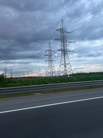 The image captures a roadside view of numerous high-voltage electricity pylons stretching across a green landscape under a cloudy sky. The pylons, made of metal lattice structures, are arranged in a line, with power lines strung between them. The foreground shows a road, likely a highway, with a white line marking its edge and a metal guardrail separating it from the embankment. Beyond the guardrail, a field of green vegetation extends towards the pylons. In the distance, a line of trees forms a backdrop, and a few construction cranes are visible, suggesting ongoing development. The sky is filled with heavy, dark clouds, creating a somber and overcast atmosphere. The scene appears to be in Zanevka, Russia, as indicated by the presence of such power infrastructure.