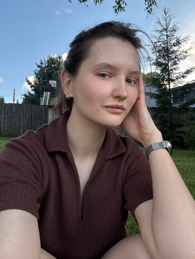 A young woman with light skin and brown hair tied back is sitting outside in a grassy area. She is wearing a brown knitted polo shirt and a silver watch on her left wrist. Her left hand is resting on her cheek, and her gaze is directed towards the camera. Behind her, a wooden fence encloses the property, with trees and a house visible in the background. The sky is blue with scattered clouds. The image appears to have been taken in the city of Burdyata, Russia.