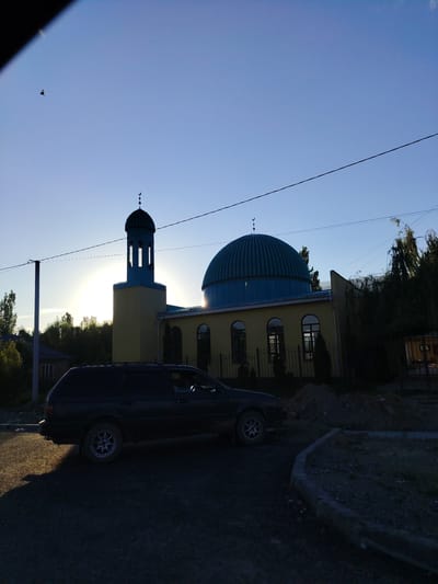 A black station wagon is parked on a gravel road in front of a mosque. The mosque is yellow with a blue dome and a blue minaret topped with a crescent moon. The sun is setting, casting a warm glow on the scene and creating lens flare. Trees and other buildings are visible in the background. The sky is clear and blue. The overall mood is peaceful and serene. There is no visible text.