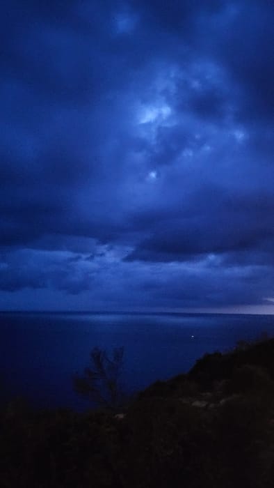 The image displays a dark, expansive seascape captured from an elevated coastal vantage point in Santa Ponça, Spain, consistent with late dusk or night conditions. The upper two-thirds of the frame are dominated by a cloudy, deep indigo-blue sky, featuring brighter, diffused light patches illuminating portions of the cloud cover. A distinct, horizontal horizon line separates the sky from the calm, dark blue expanse of the sea. A single, sharp point of light, possibly from a distant vessel or coastal feature, is visible on the water's surface in the middle-ground, slightly right of center. The lower third of the image reveals a dark, indistinct landmass, likely a rocky hillside or cliff, with sparse, silhouetted vegetation, including a few prominent dark tree or shrub outlines extending into the left side of the frame. No individuals, specific actions, or interactions are discernible.