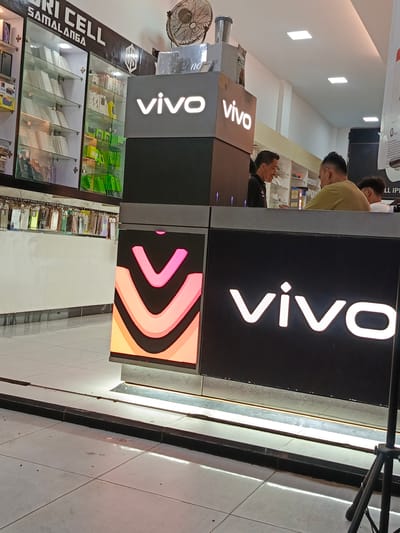 The image depicts the interior of a Vivo mobile phone store in Namplohbaro, Indonesia. In the foreground, a large, illuminated Vivo logo is visible on a black counter. To the left of the logo, a display panel shows a vibrant gradient of pink and orange curved shapes. Behind the counter, two men are interacting. One man, wearing a black shirt, appears to be an employee and is facing the other man, who is wearing a light brown t-shirt. The man in the light brown shirt is facing away from the camera. Another man, partially visible, is standing behind him. Behind the counter, shelves display various mobile phone accessories, including phone cases and boxes, with some items in green packaging. To the far left, a glass display cabinet showcases more phone cases. The store appears to be well-lit, with recessed lighting in the ceiling. A vintage fan is mounted on the wall above the display cabinets. The floor is tiled with light-colored square tiles.
