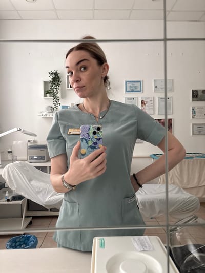 A woman in light green scrubs is taking a selfie in a mirror. She is wearing a watch on her left wrist, a beaded bracelet on her right wrist, and a silver necklace. Her phone has a blue, purple, and yellow floral case. Behind her, on a white wall, are various framed certificates and a small plant. A white cosmetic device and various pieces of medical equipment are visible, including what appears to be an autoclave. A medical table or chair covered in white plastic sheeting is also visible. The location is potentially in Rahya, Russia.
