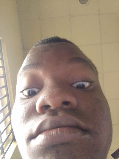 A close-up, low-angle selfie captures the face of a young person of African descent, filling the frame. The subject's eyes are wide and looking directly at the camera. Their nose is prominent, and their lips are slightly parted. The skin texture is visible, with some pores and a slight sheen. The background appears to be an indoor ceiling with tiled panels and a circular light fixture. To the left of the subject, a barred window with vertical and horizontal elements is partially visible, suggesting an interior space with an external view. The lighting is bright, casting some shadows. The image is grainy, indicating it was taken with a low-resolution camera or in less than ideal lighting conditions. The subject's head is tilted, and the perspective is distorted due to the close proximity of the camera and the wide-angle lens effect. The location is identified as Eleweran, Nigeria, suggesting an urban or semi-urban environment.