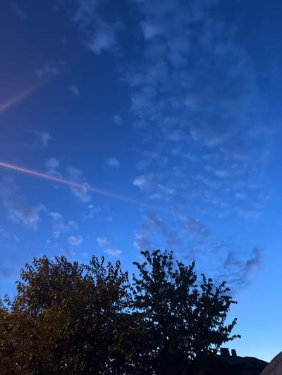 The image captures a twilight sky over Topolchane, Bulgaria, as dusk settles. The primary subject is the expansive blue sky, dotted with scattered, wispy clouds. A prominent feature is a thin, almost ethereal band of light that diagonally crosses the frame from bottom left to top right, possibly a contrail or a reflection.
On the right side of the image, the dark silhouette of a deciduous tree fills a significant portion of the frame. Its leaves are dense and show hints of golden light filtering through, suggesting artificial illumination from below or from a nearby source. Above the tree, a dark, architectural element is visible, hinting at a building or structure, with two chimney-like features protruding.
The time of day is clearly evening or dusk, given the deep blue of the sky and the fading light. The weather appears calm, with the clouds being light and dispersed. There are no people or specific objects that are the main focus, beyond the natural elements of the sky and the tree, and the hint of human-made structures. The overall mood is serene and contemplative, characteristic of the transition from day to night. No visible text is present in the image.