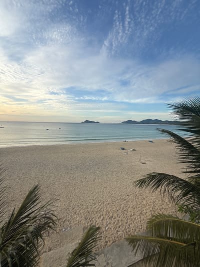 The image depicts a serene beach scene in Lệ Uyên, Vietnam, bathed in the soft light of either dawn or dusk. The foreground is dominated by the sandy expanse of the beach, with some subtle textures and shadows indicating it's a natural, untamed environment. Palm fronds frame the scene from the top and right, suggesting a tropical locale. In the middle ground, the calm, turquoise ocean meets the shore with gentle waves. Several small boats are visible scattered on the water, hinting at local fishing activities or leisure. The horizon is marked by a chain of mountainous islands, receding into the distance and softened by atmospheric haze. The sky above is a canvas of blue, with wispy white clouds creating a dynamic yet peaceful atmosphere. There are no people prominently featured in the image, contributing to the tranquil and unpopulated feel of the beach. The overall impression is one of natural beauty and quiet solitude.