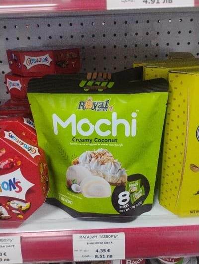 The scene depicts a close-up view of a retail shelf inside "МАГАЗИН "ИЗВОРЪ"" in Byal Izvor, Bulgaria. The central subject is a lime green stand-up pouch of "Royal Family Mochi" with "Creamy Coconut" flavor, stating it contains 8 pieces and has a net weight of 120g, originating from Taiwan. The pouch design includes an image of mochi, whipped cream, and a half coconut. To the immediate left, a red hexagonal box of "Celebrations" chocolates is partially visible, with a second identical box stacked beneath it. To the right, a portion of a tall, yellow rectangular package is present. All products are displayed on a white shelf. Above this shelf, a grey perforated metal pegboard is visible. Below the white shelf, a red horizontal strip features a price label for "Б-НИ МОЧИ 120 ГР." (Mochi 120 gr.), indicating a price of 4.35 € and 8.51 ЛВ. No individuals are visible in the image.