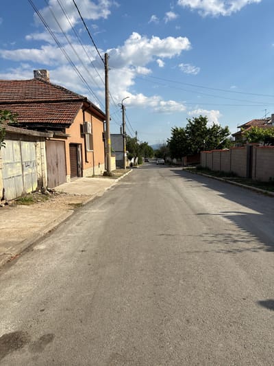 A street scene in Kran, Bulgaria, shows a paved road lined with houses and fences under a bright blue sky with scattered white clouds. The road curves slightly into the distance. On the left side of the road, there is a house with a red tile roof, a light orange facade, and a garage with two large metal doors, one of which is marked with graffiti. To the right of the garage is a smaller brown door. Further down this side of the street, a modern grey house is visible. On the right side of the road, there is a long, grey brick fence with red caps. Trees and other houses are visible beyond the fence. Two cars are parked on the road in the distance. Electrical poles and wires are present on both sides of the street. The lighting suggests it is daytime, likely late morning or afternoon, given the shadows.