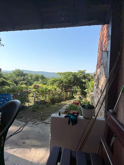 A sunny outdoor scene, viewed from under a dark overhang, looking out onto a lush garden with rolling hills in the distance. In the foreground, a dark chair is partially visible, and a blue barrel with white polka dots sits to the left. The garden features a variety of plants, including blooming pink roses and a large, leafy tree. A stone wall and a portion of a brick building are visible on the right side of the frame. Gardening tools, including a pair of blue and black gloves and a white pot with a plant, are placed on a ledge. The sky is clear and blue, and the overall impression is one of a peaceful, rural setting in Gorno Pavlikene, Bulgaria.